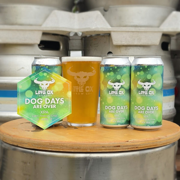 Cans of Little Ox Brewery Dog Days Are Over XPA with a pint full of beer on a background of kegs.