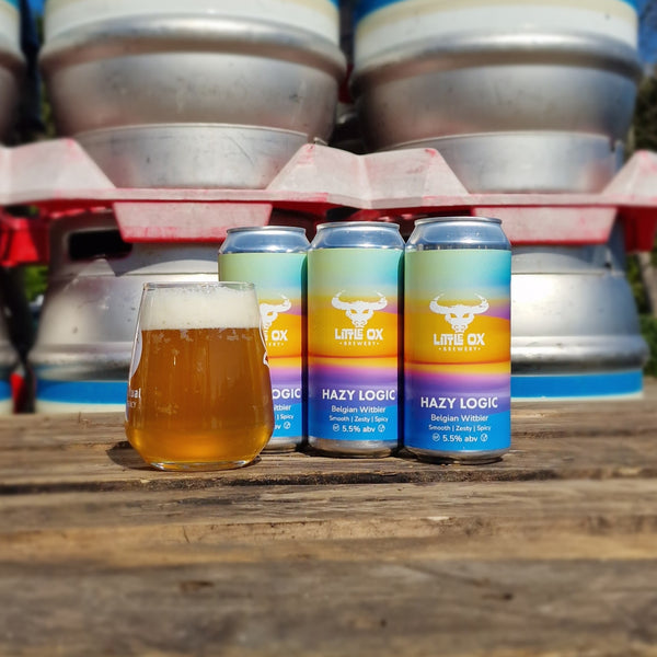 Cans of Little ox Hazy Logic beer with a glass of beer on a wooden surface, surrounded by metal kegs.   
