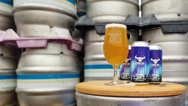 Lumen cans and glass of beer on a wooden table with keg stack behind.