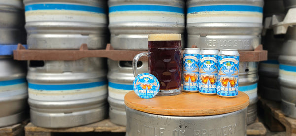 Three cans of Little Ox Brewery Stein Alive Munich Dunkel 4.5%, with a stein full of beer and a beer badge on a table in front of steel beer kegs.