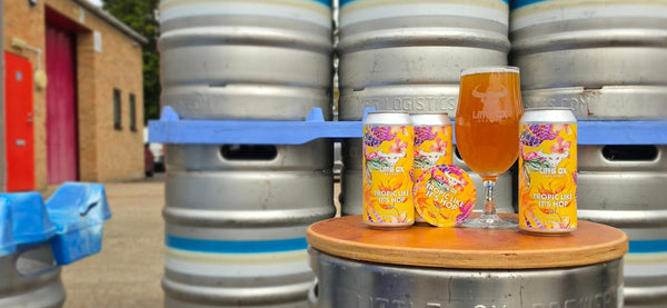 Three cans of Little Ox Brewery Tropic Like It's Hop DIPA with a glass of beer displayed in front of stacked stainless steel kegs.