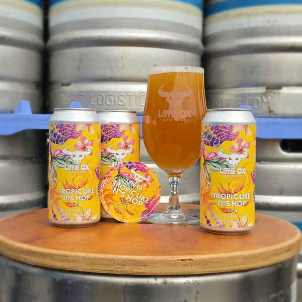 Three cans of Little Ox Brewery 'Tropic Like It's Hop' beer, including two labeled cans and a glass of beer, arranged on a  barrel with stacked kegs in the background.